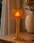 An amber glass candlestick table lamp with a lit candle inside, styled on a wooden table next to a beige ceramic vase with greenery.