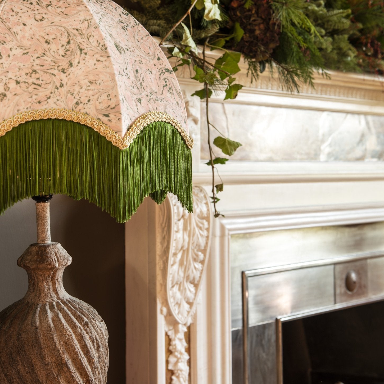 A close-up of a decorative table lamp with a patterned beige and green damask fabric shade, a green fringe trim, and a textured stone base, positioned next to an ornate fireplace mantel with greenery.