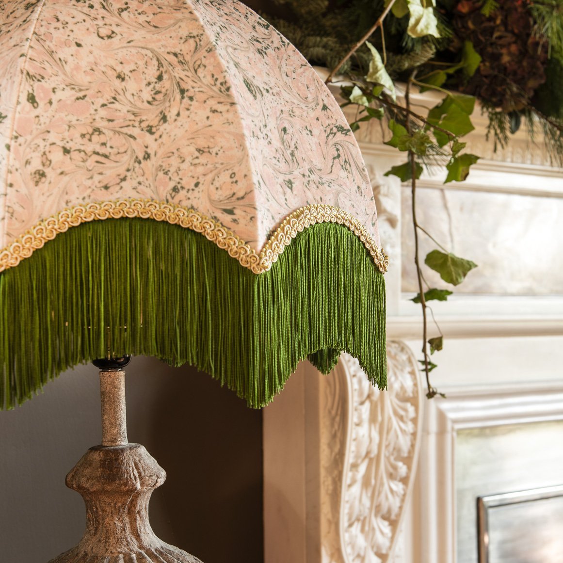 A close-up of an ornate lamp shade with a pale blush pink and green floral pattern, a gold decorative trim, and a long green fringe, mounted on a textured stone-like lamp base in an indoor setting.
