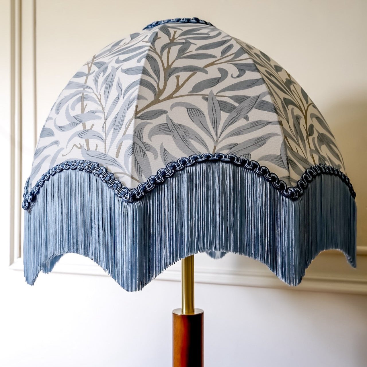 A dome-shaped table lamp shade with a blue and gray botanical leaf pattern, finished with a scalloped fringe trim and a gold-toned metal stem.