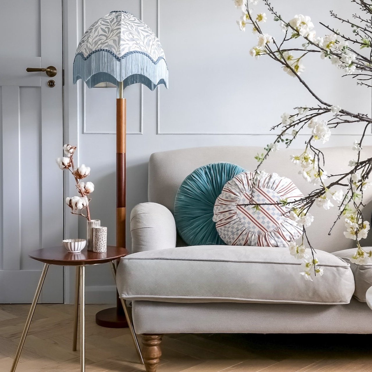 A styled interior scene with a light beige armchair, patterned throw pillows, a floor lamp with a blue-and-white scalloped shade, a small round side table, and flowering branches in the foreground.