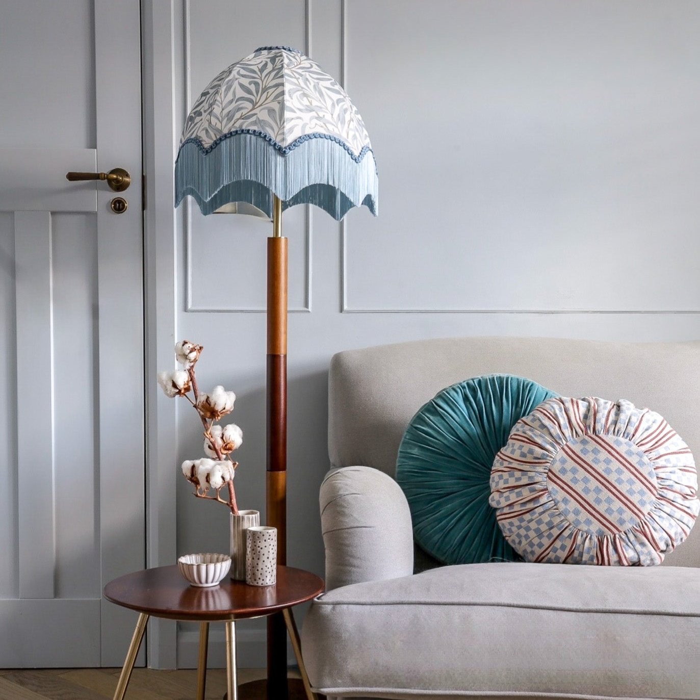 A tall floor lamp with a patterned white and blue shade, a light blue ruffled trim, and a multi-toned wooden pole stands beside a light gray sofa with decorative pillows in a styled living room.