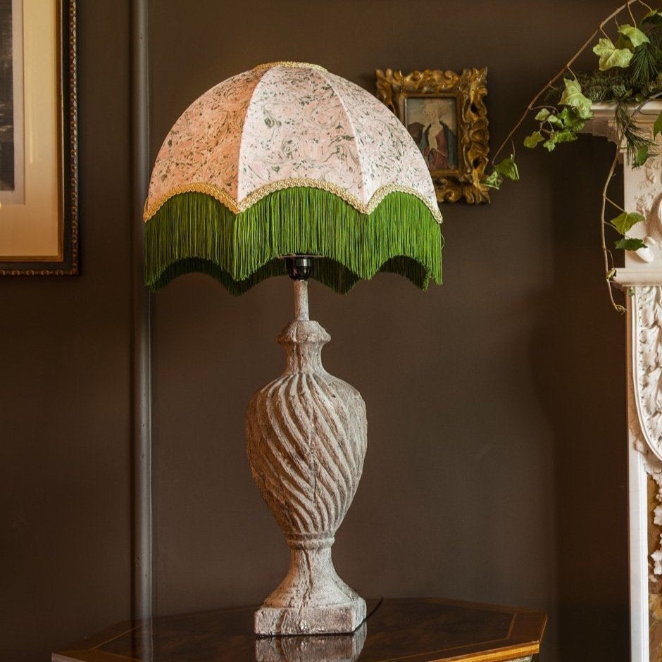 An ornate table lamp with a distressed white textured urn-shaped base and a dome-shaped shade featuring a beige and white floral pattern, green fringe trim, and a black band. The lamp is styled on a dark wooden table against a dark brown wall with framed art and greenery in the background.
