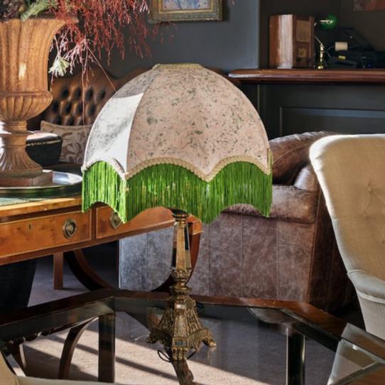 An ornate table lamp with a dome-shaped beige floral fabric shade and bright green fringe trim, set on a glass-top table in a furnished room.