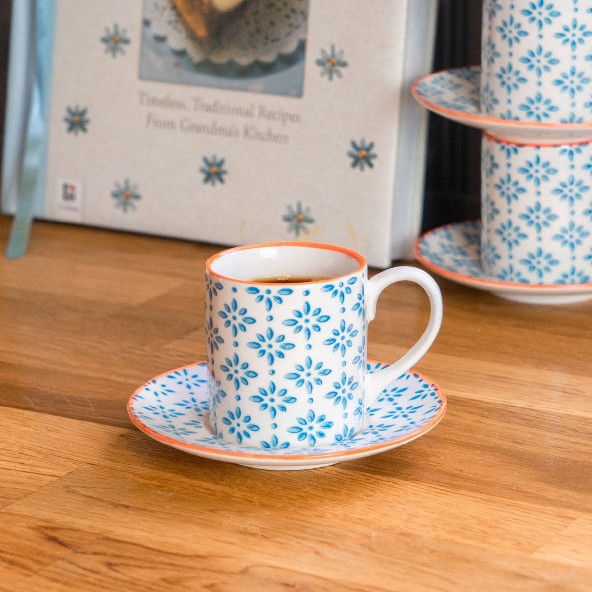 A white ceramic coffee cup with a blue floral pattern and orange rim sits on a matching saucer on a wooden table, with additional matching cups and a recipe book in the background.