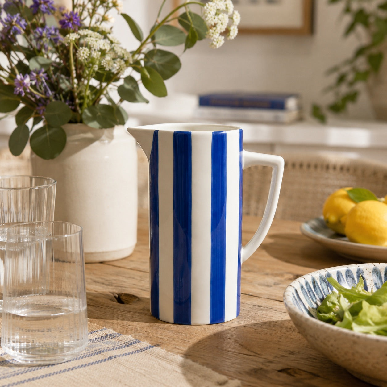 A white ceramic pitcher with bold vertical blue stripes sits on a wooden table in a bright kitchen setting, surrounded by glassware, flowers, lemons, and a bowl of salad.