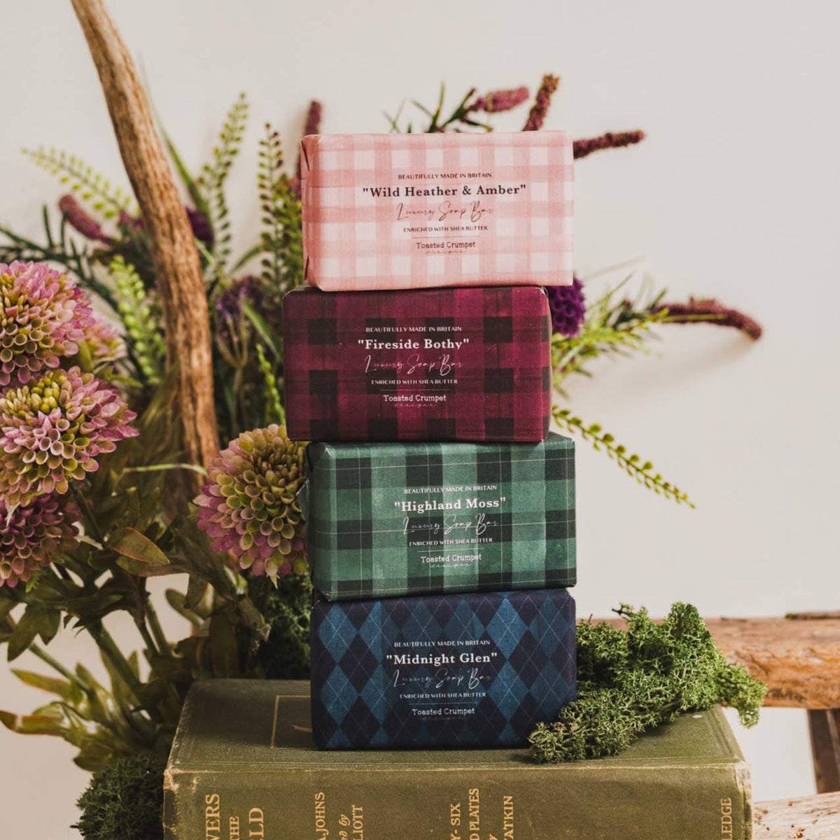 Wild Heather &amp; Amber in pink gingham, Fireside Bothy in burgundy and black plaid, Highland Moss in green and black plaid, and Midnight Glen in navy blue diamond-patterned packaging