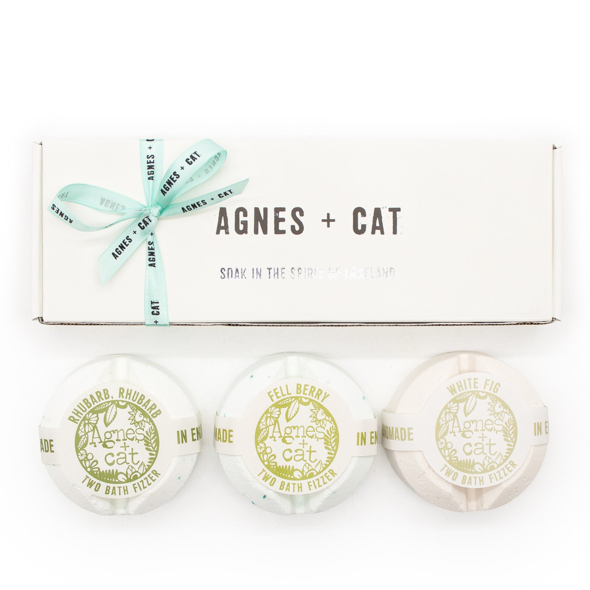 Three round bath fizzles with pale green and white labels are displayed in front of their white gift box, which is tied with a mint green ribbon. The box features the brand name 'AGNES + CAT'.