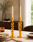 A pair of amber glass candlestick holders with stacked round beads and white taper candles, set on a dining table in a softly lit room.