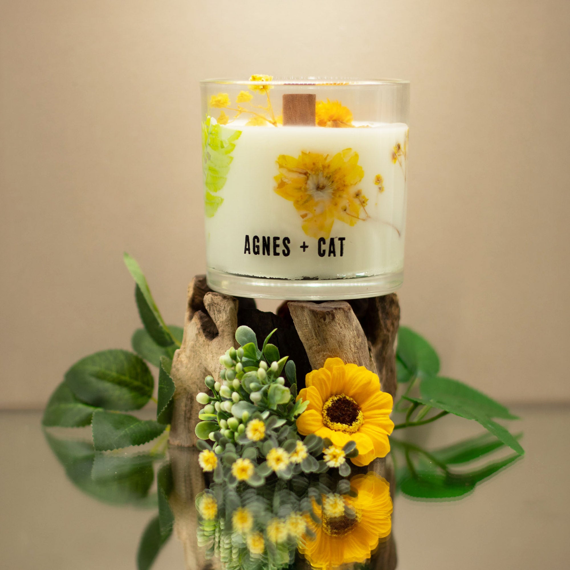 Candle with floral design and 'AGNES + CAT' branding on a wooden stand with plants.