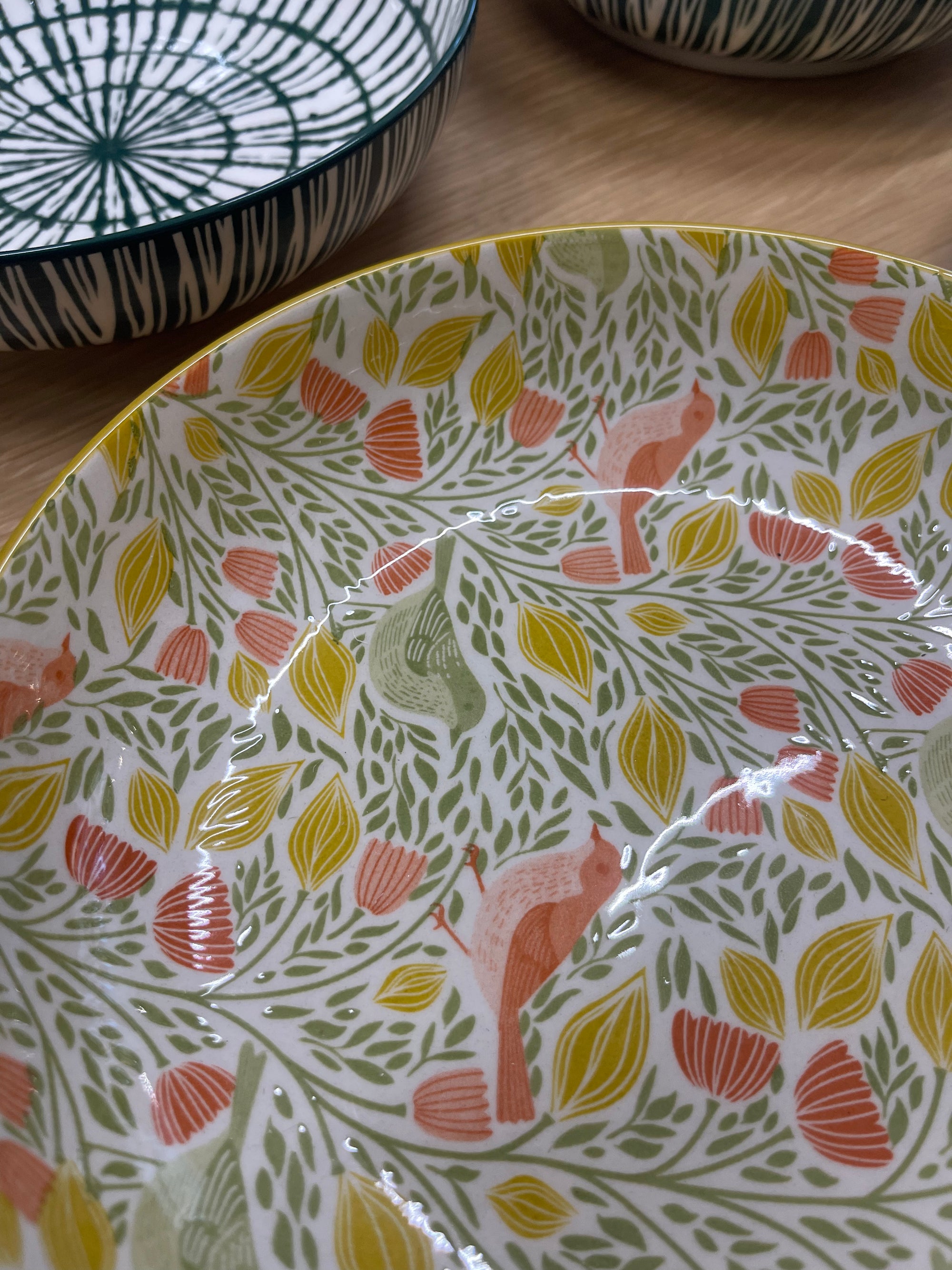 A close-up of a ceramic bowl with a white base decorated with a dense pattern of green leaves, yellow and orange tulips, and pink birds. The bowl is shown on a wooden surface, with other bowls partially visible in the background.