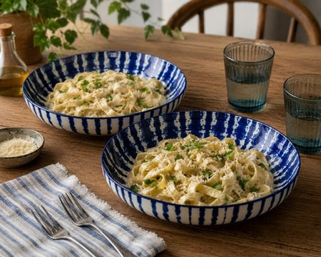 Two blue and white striped ceramic bowls filled with creamy pasta on a wooden dining table, with blue-tinted glasses, a striped napkin, forks, and a small dish of grated cheese nearby.