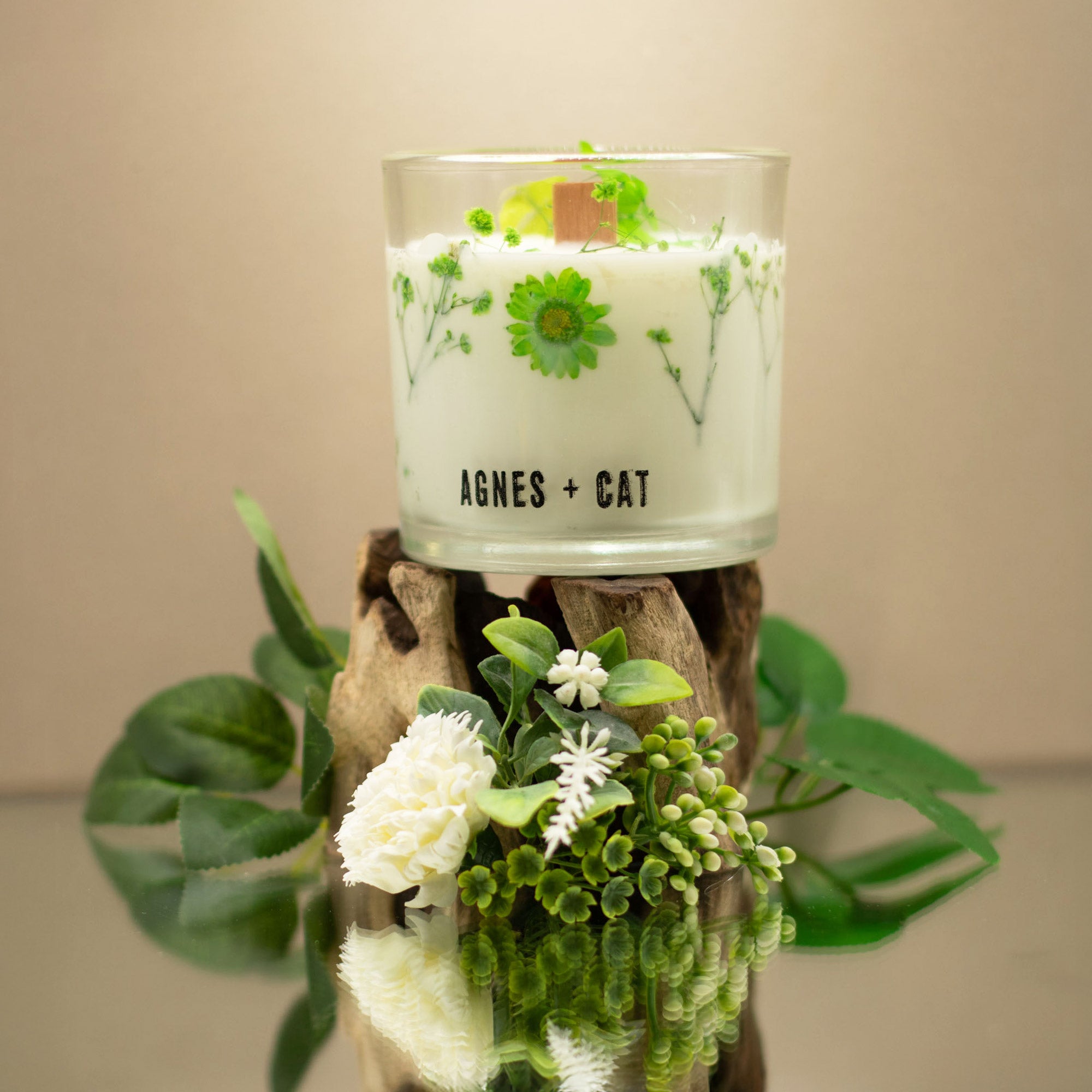 Candle with floral design and 'AGNES + CAT' branding on a wooden stand with greenery.