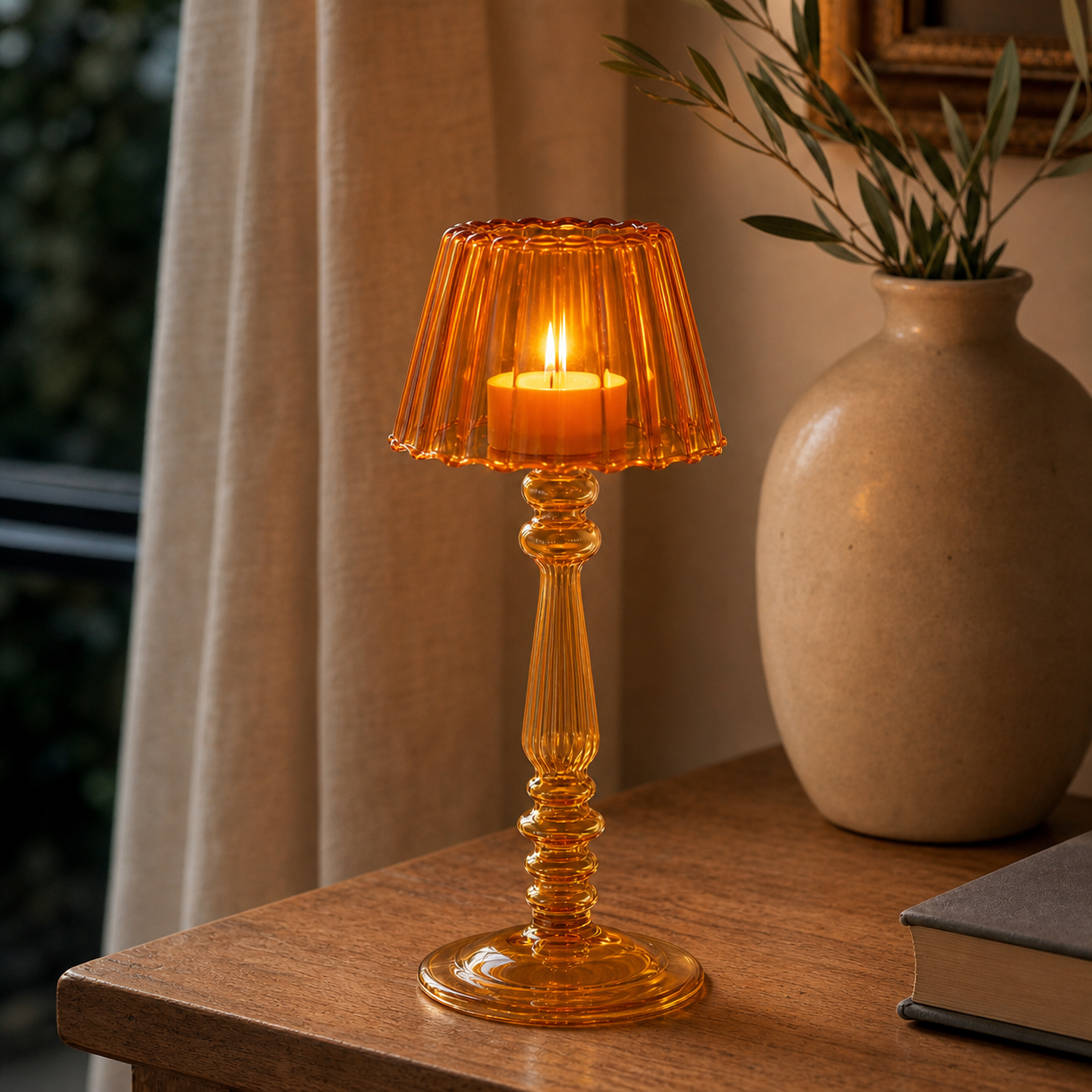 An amber glass candlestick table lamp with a lit candle inside, styled on a wooden table next to a beige ceramic vase with greenery.