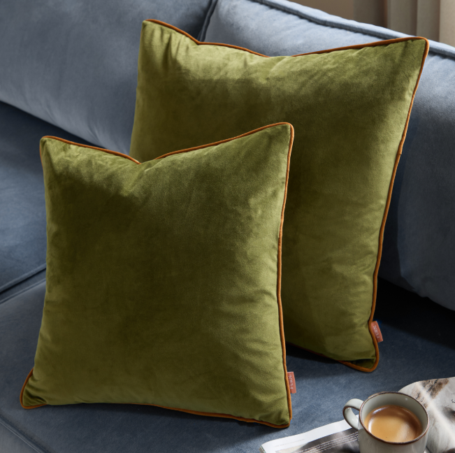 Two green velvet pillows with orange piping on a gray sofa.