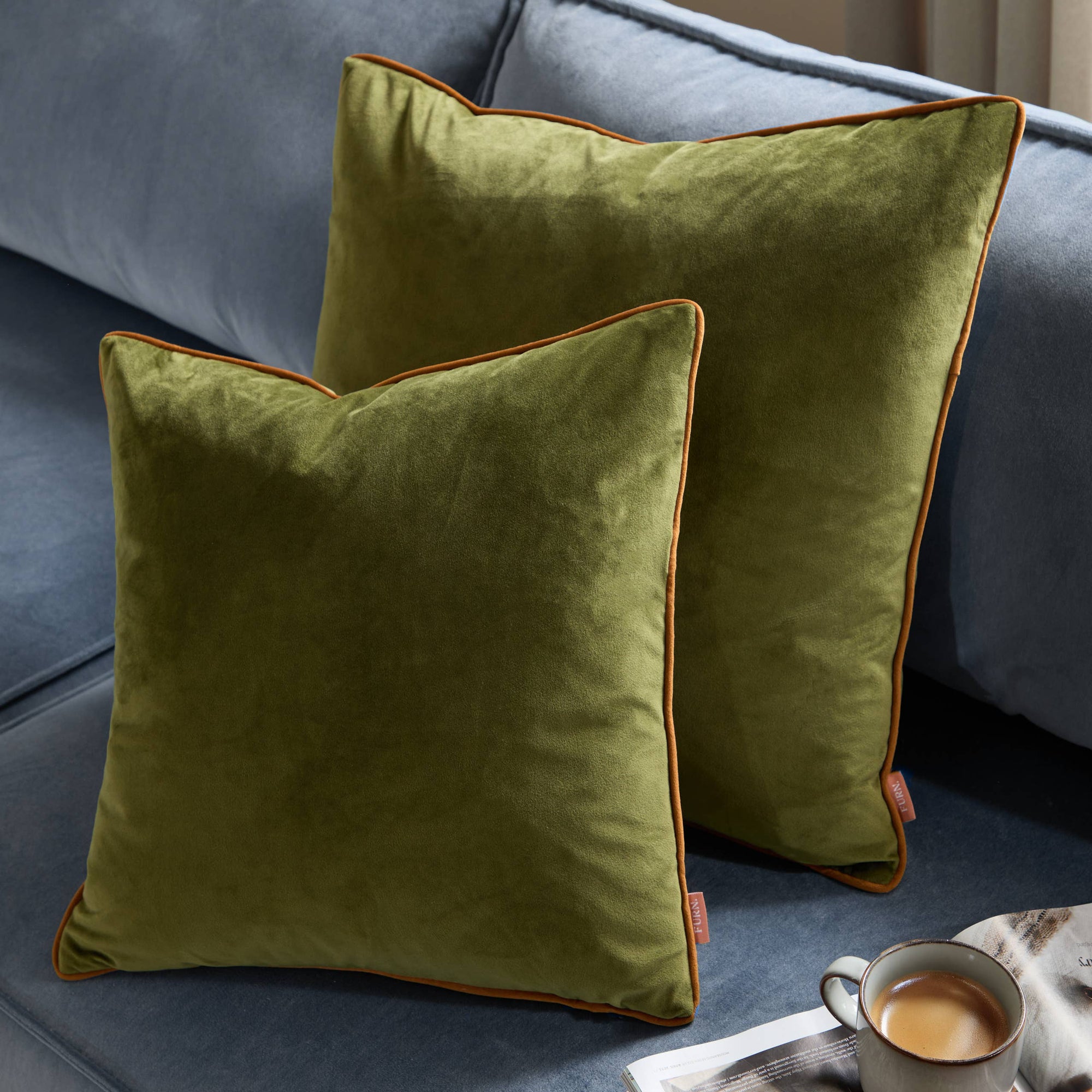 Two green velvet pillows with orange piping on a gray sofa.