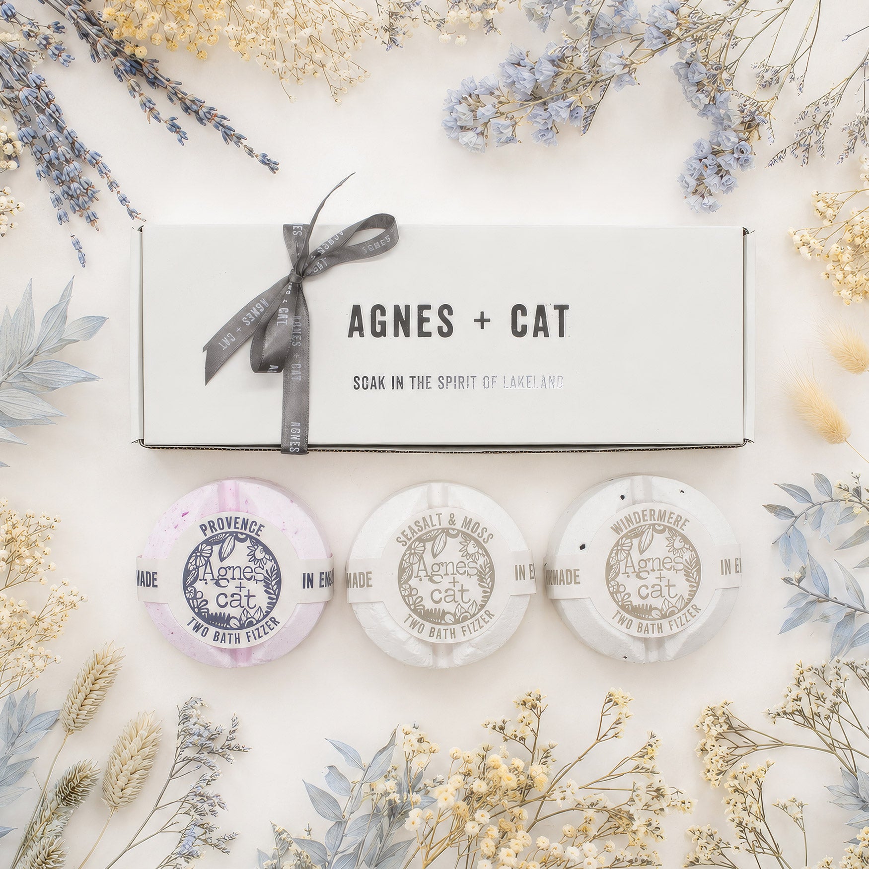 A boxed bath soak product labeled 'AGNES + CAT' with a gray ribbon, shown on a light neutral background surrounded by dried flowers. Three round wrapped bath salts or pads are displayed below the box.