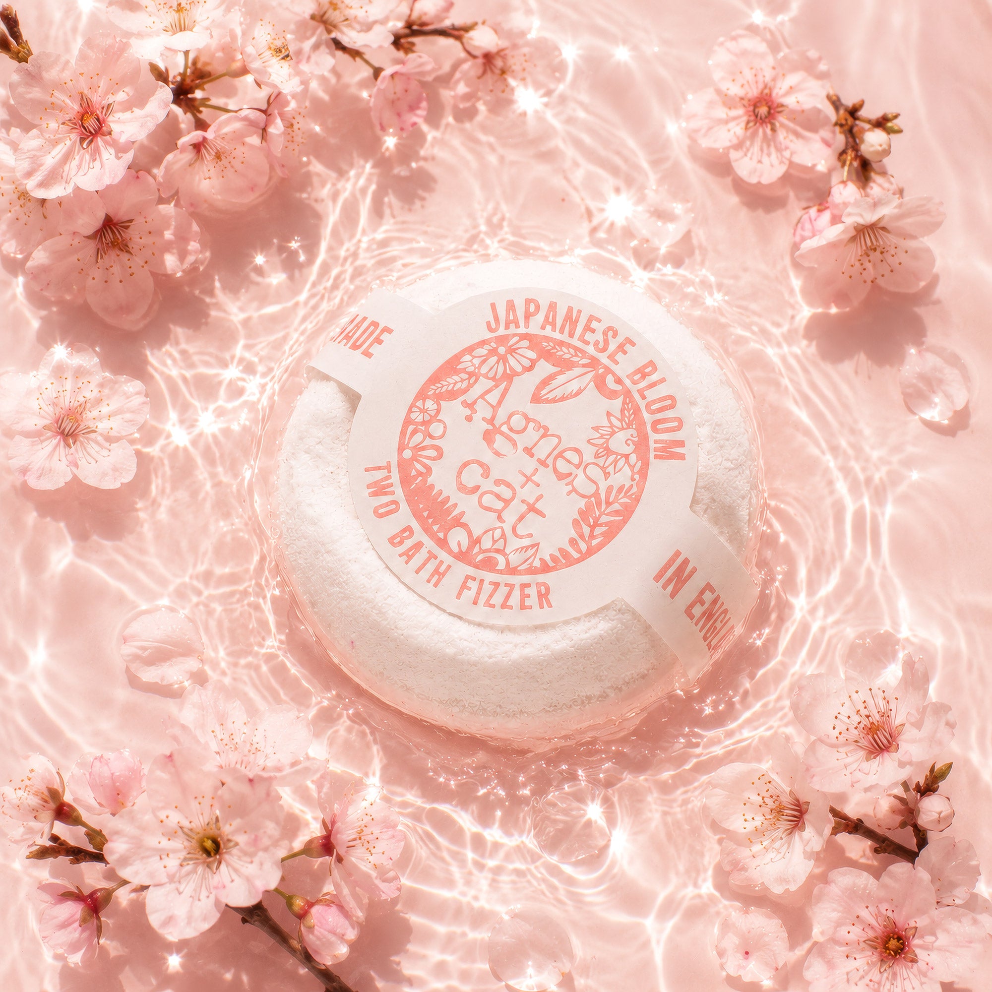 A round white bath fizzer product labeled 'Hono Cat Japanese Bloom Two Bath Fizzer' placed on a pink water surface surrounded by pink cherry blossoms.