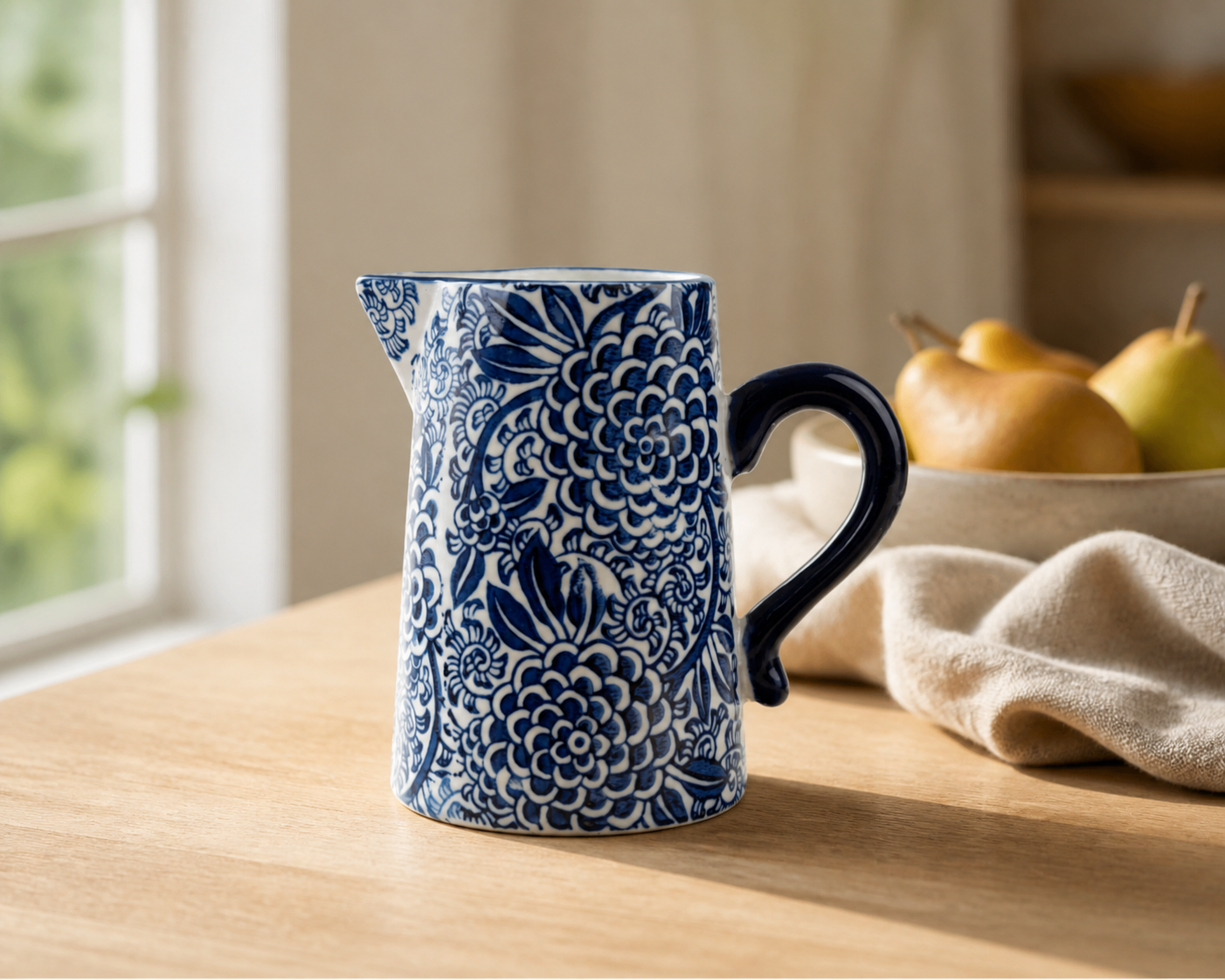 A blue and white floral patterned ceramic pitcher with a dark blue handle, sitting on a wooden table in a kitchen setting with pears and a cloth in the background.