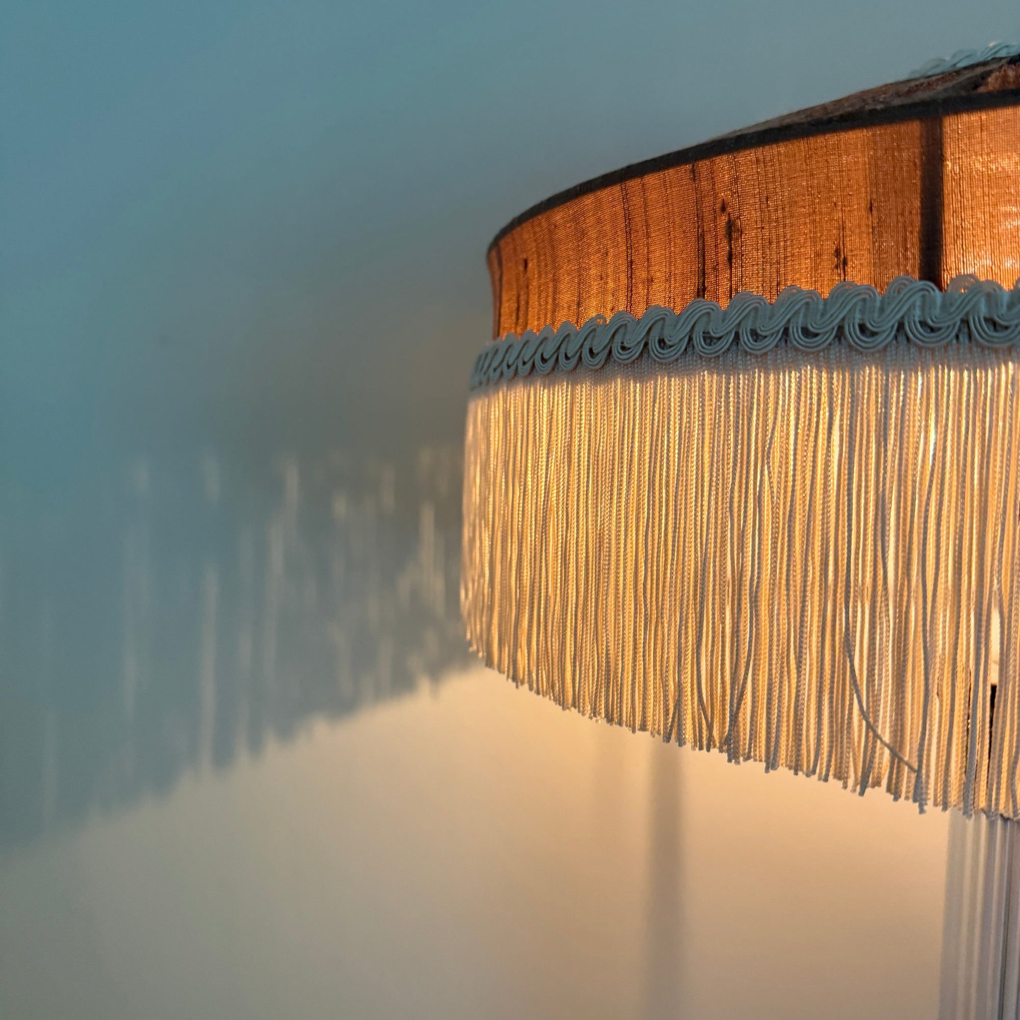 A close-up of a lamp shade with a warm brown wood-look top band, a light blue scalloped fringe trim, and long cream fringe hanging below, casting a soft shadow against a pale wall.