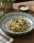 A wide, shallow ceramic bowl with a black and white striped pattern, filled with pasta and garnished with basil leaves and parmesan cheese, set on a wooden dining table.