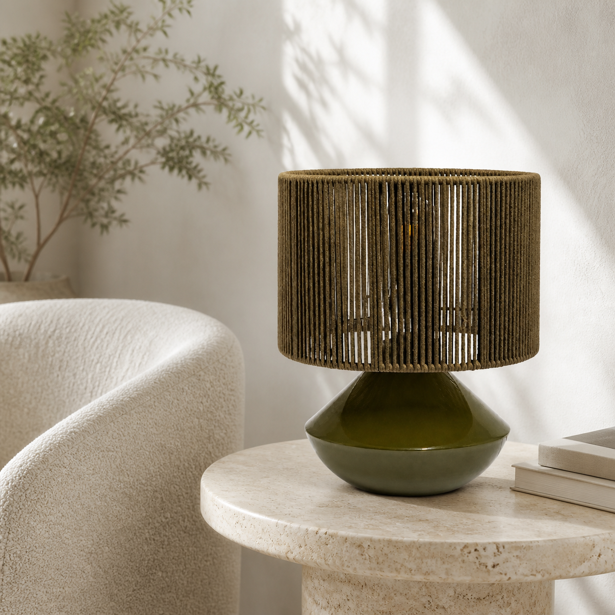 A table lamp with a dark olive green ceramic base and a cylindrical woven shade in a warm brown tone, styled on a round stone side table in a bright, modern room.