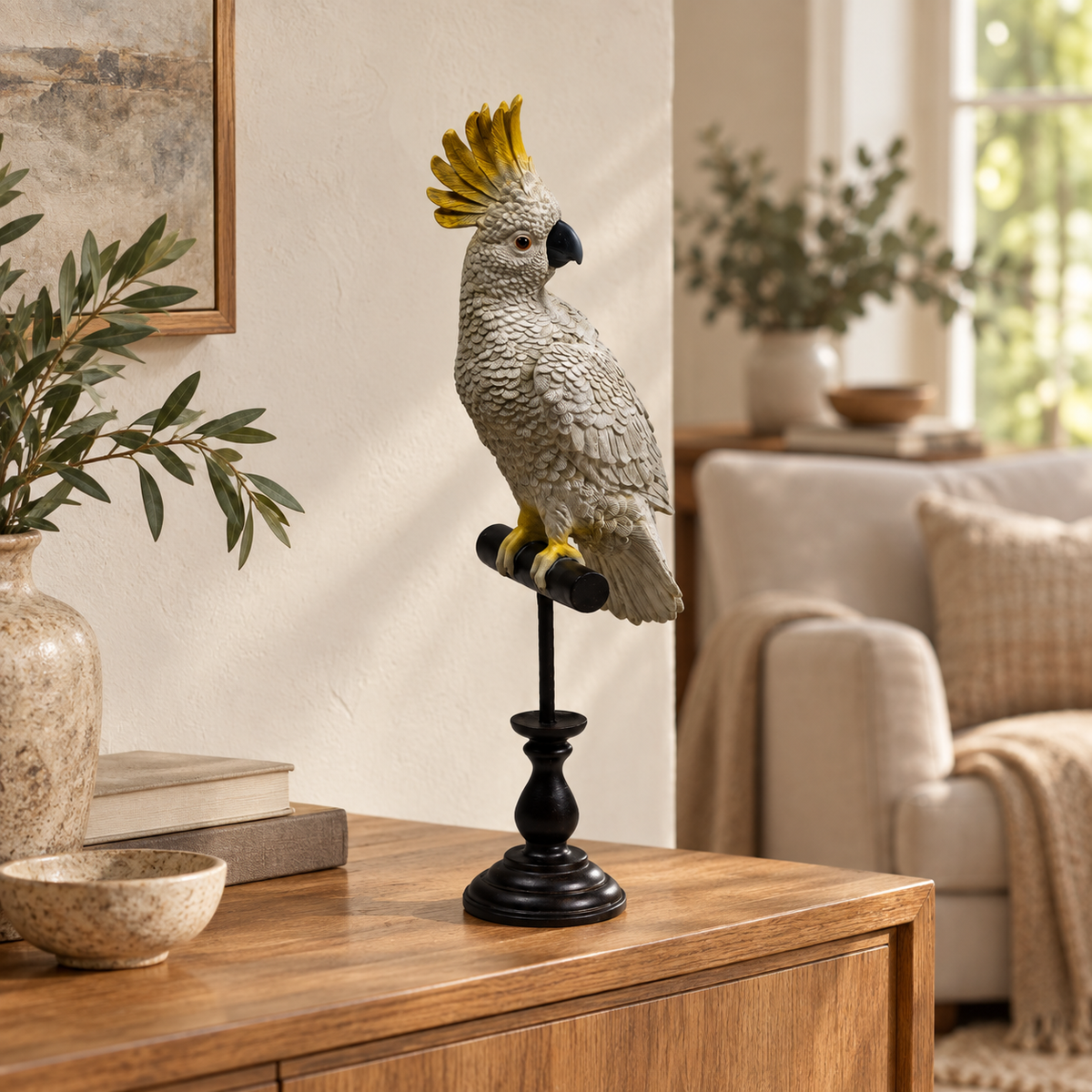 A decorative grey cockatoo figurine with a bright yellow crest perched on a black stand, displayed on a wooden console table in a styled living room setting.