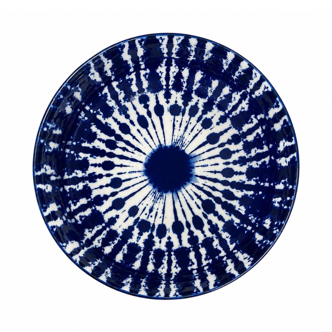 A round ceramic plate featuring a bold radial tie-dye or ink-blot pattern in deep cobalt blue and white, photographed from directly above on a solid white background.