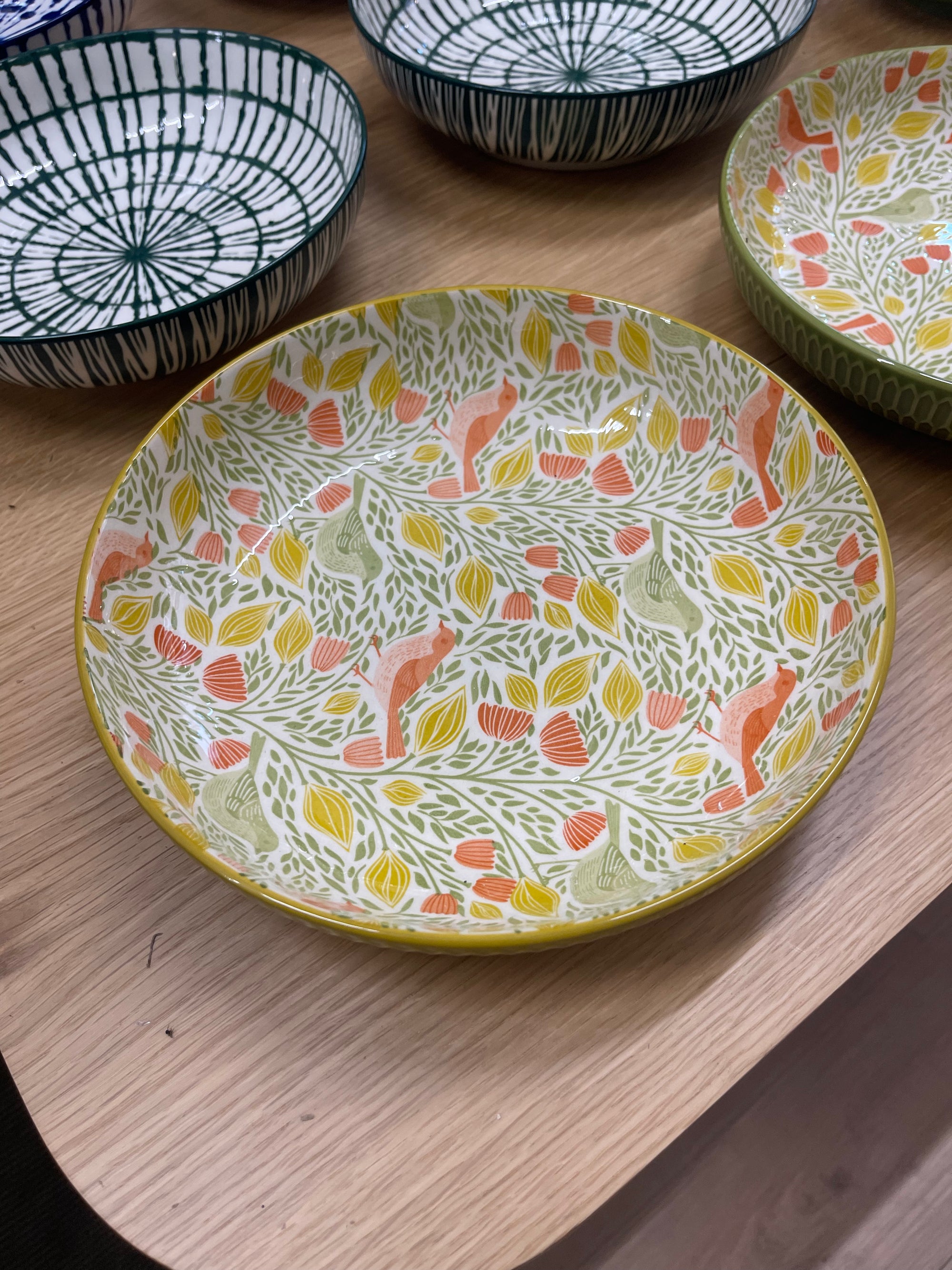 A round ceramic plate with a white base decorated with a colorful leaf and bird pattern in yellow, orange, and green, sitting on a light wood table with other patterned plates in the background.