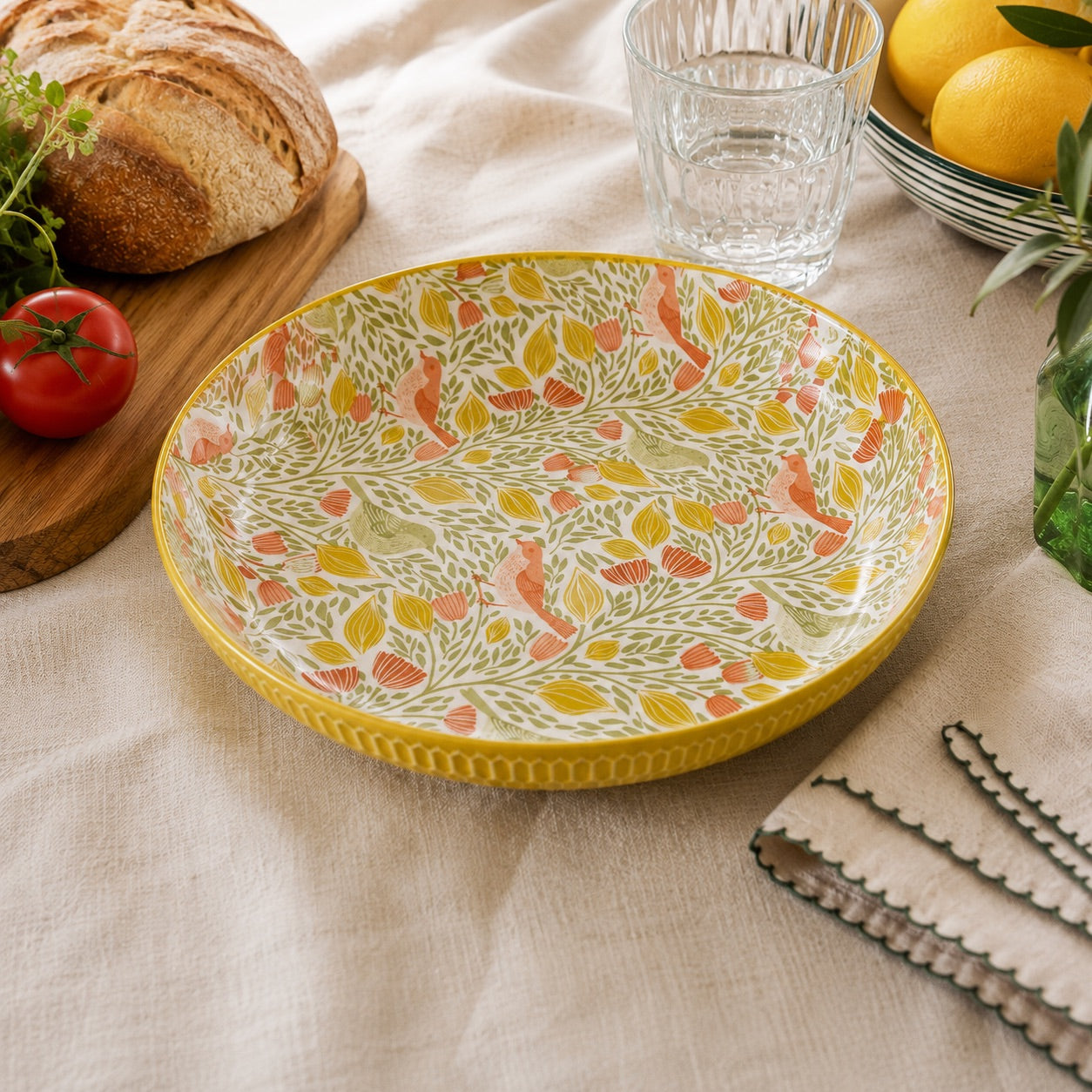 A round ceramic serving platter with a yellow rim and a white interior decorated with a botanical pattern of yellow, orange, and green leaves, styled on a beige linen tablecloth with a loaf of bread, tomatoes, lemons, and a glass in the background.