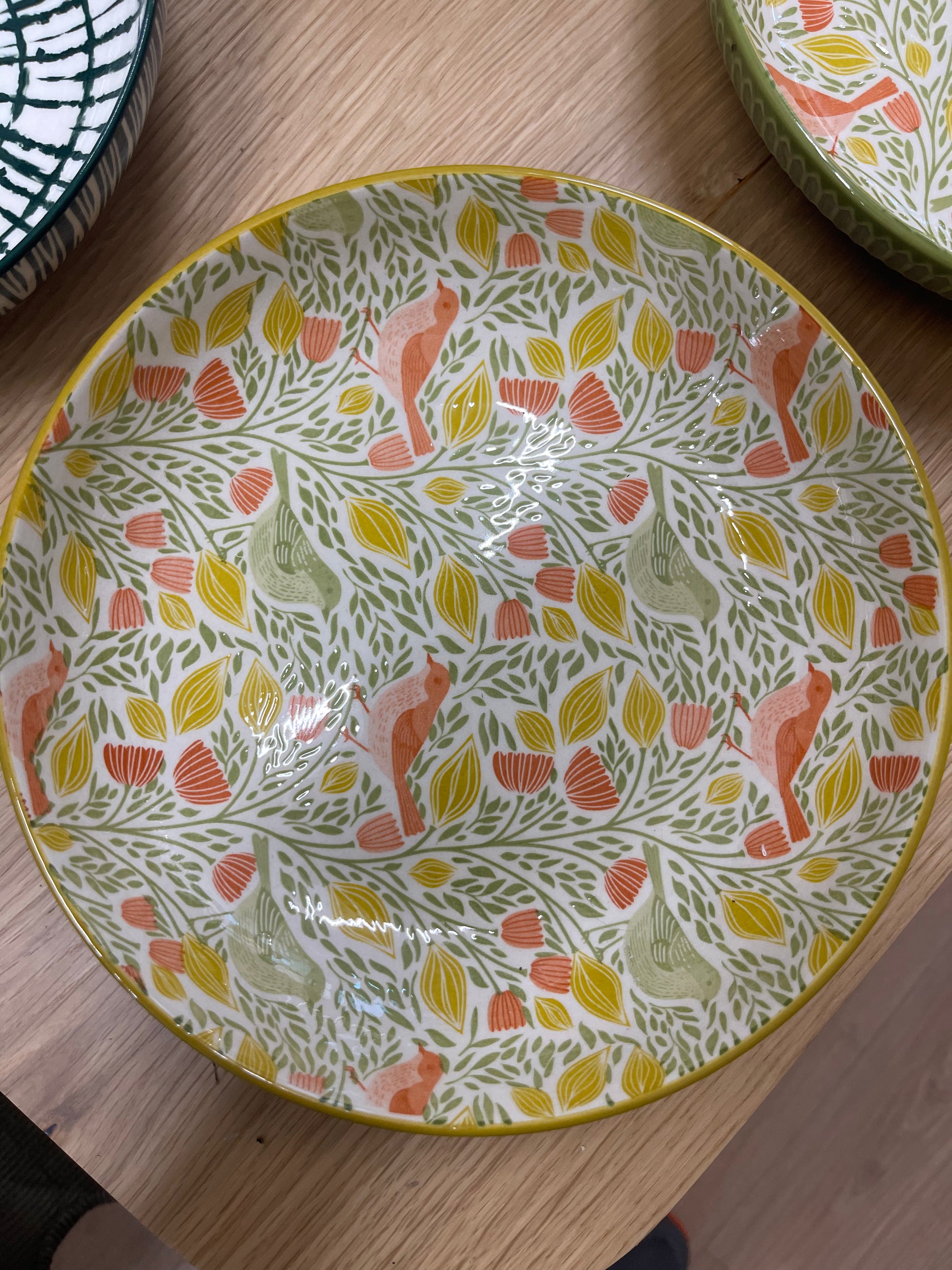 A large ceramic bowl with a white base decorated with a colorful pattern of green leaves, yellow and orange leaves, and orange birds, shown on a wooden table.