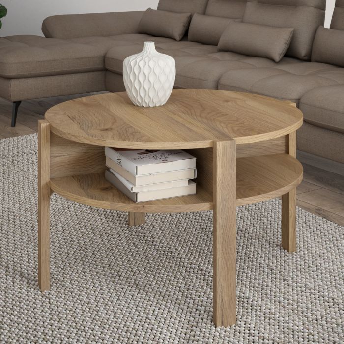 A round wooden coffee table with a natural oak finish, featuring two open shelves and styled with a white textured vase and books, set in a living room on a woven rug in front of a brown sectional sofa.