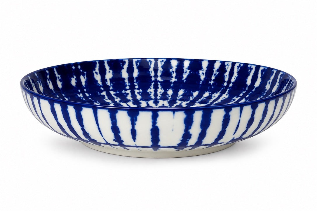 A wide, shallow ceramic bowl with a cobalt blue and white vertical tie-dye pattern, shown on a clean white background.