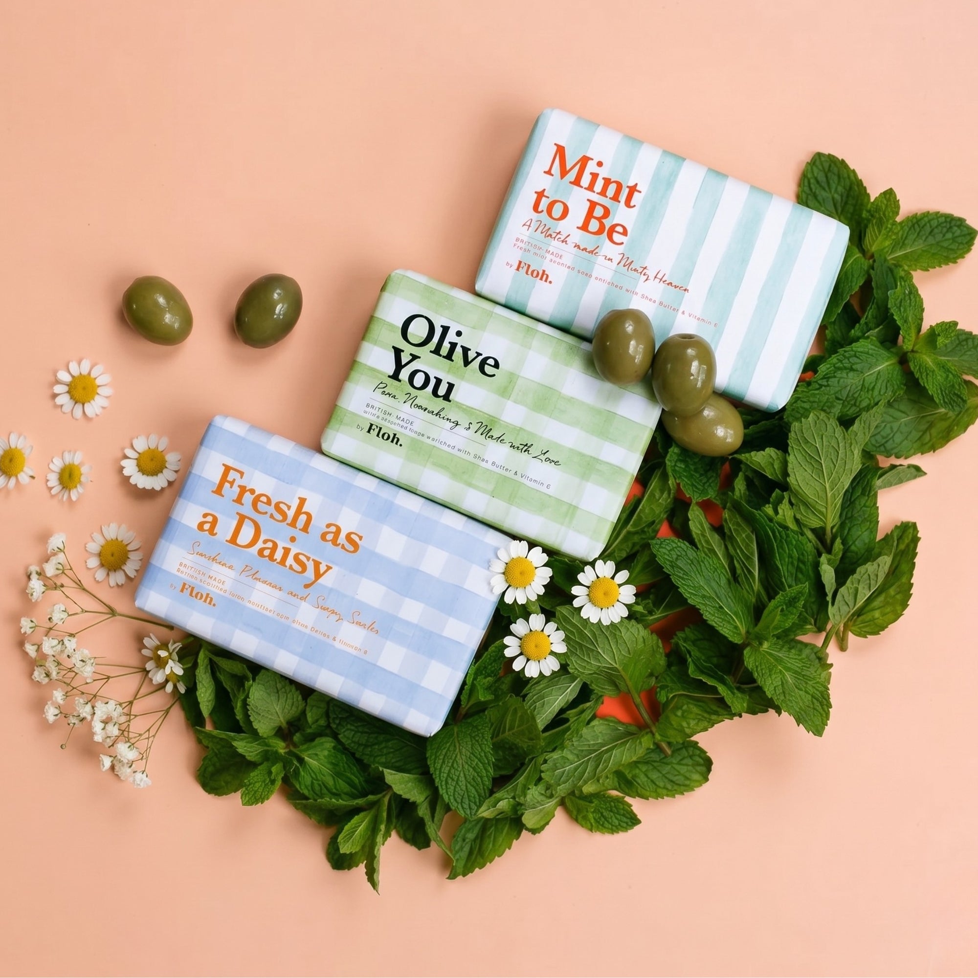 Three soap bars with decorative packaging on a pink background with green leaves and flowers.