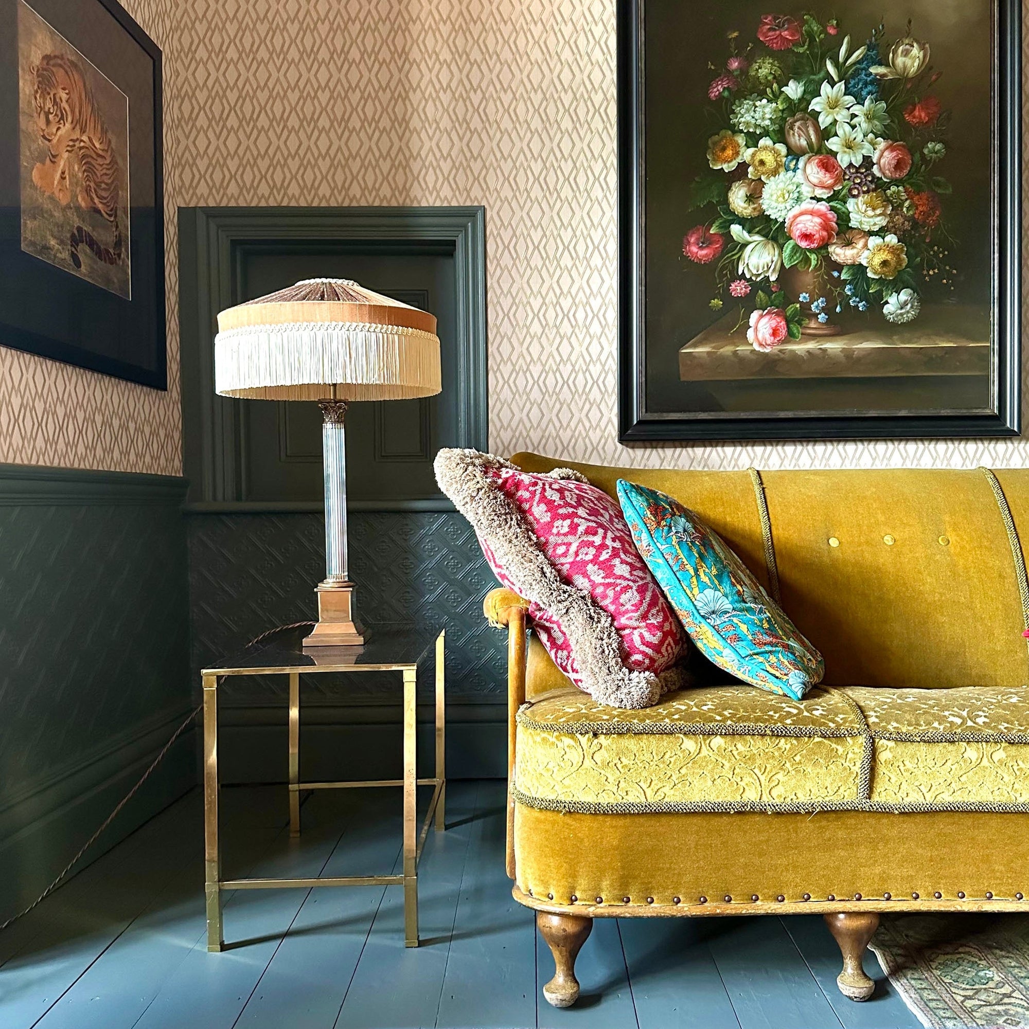 A styled interior scene featuring a mustard yellow velvet sofa with decorative throw pillows, a table lamp on a small brass side table, patterned wallpaper, dark paneled wainscoting, and framed floral wall art.