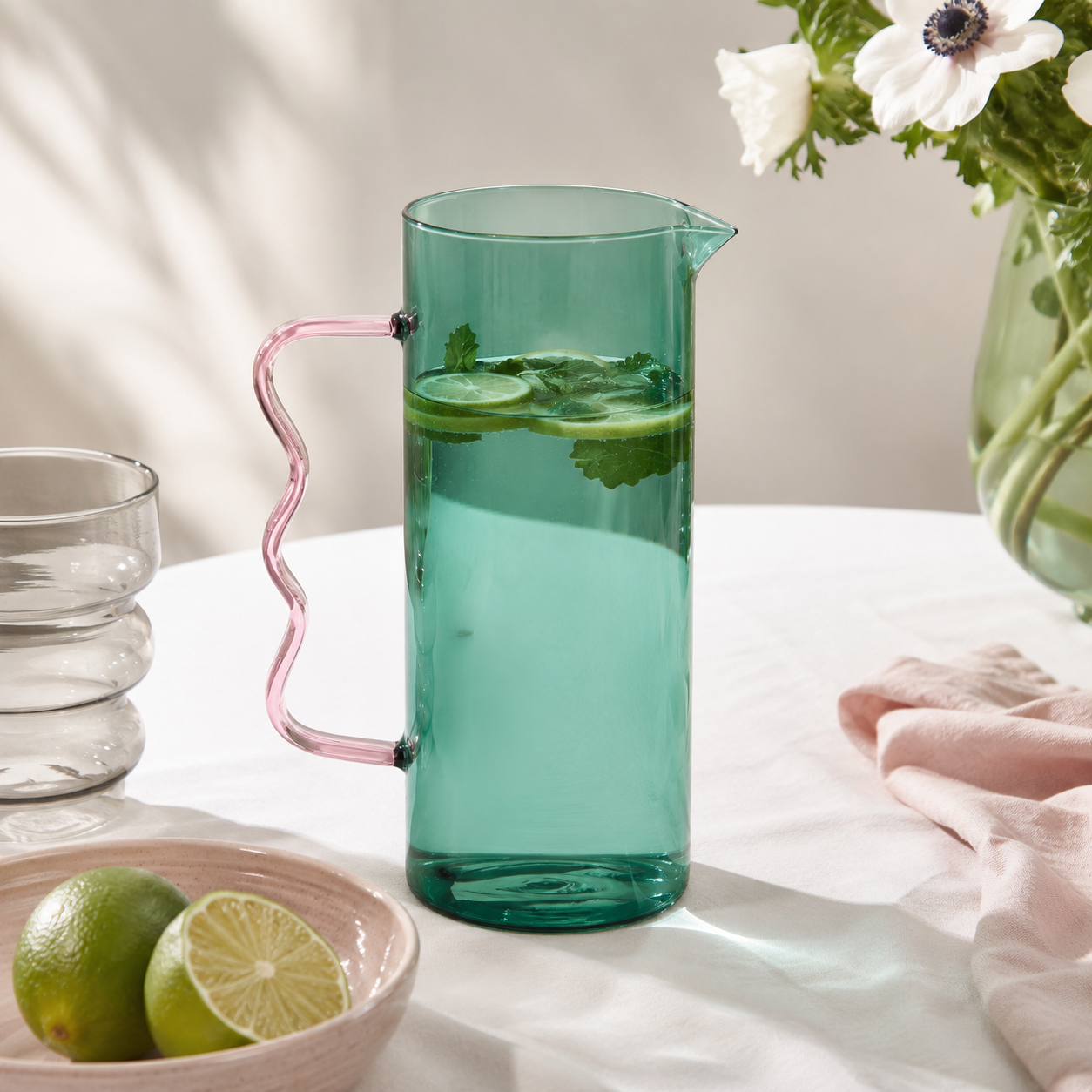 Green glass pitcher with pink handle filled with water and lime slices on a white surface.