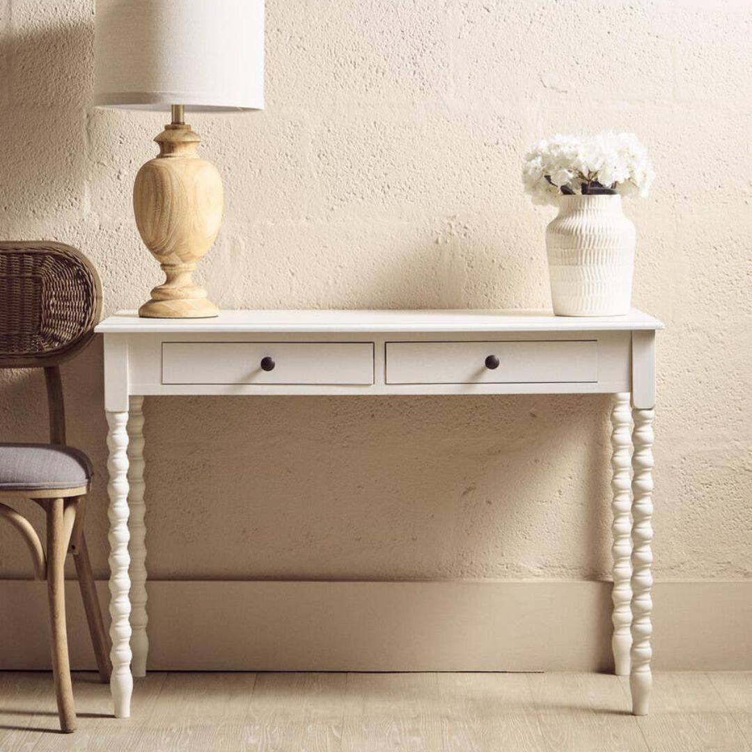 White console table with two drawers, a lamp, and a vase against a beige wall.