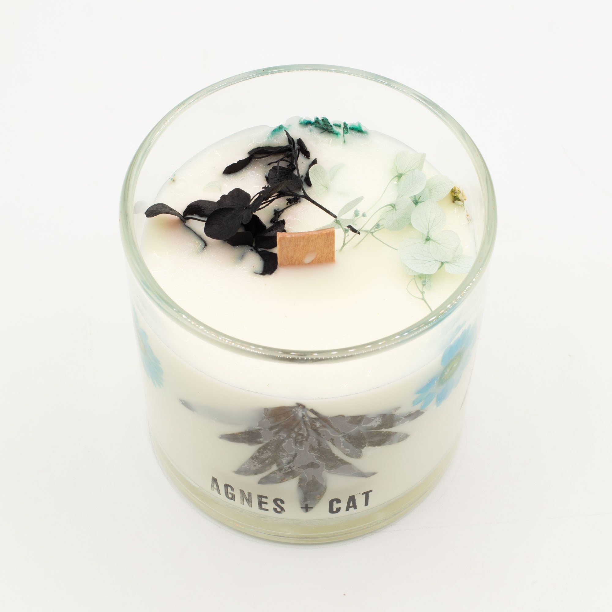 A glass jar candle with white wax, decorated with dried black flowers, pale green petals, and a small brown cube, on a white background. The glass features the text 'AGNES CAT' and a botanical illustration.
